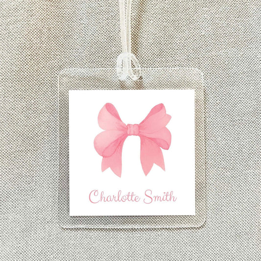 A clear name tag with a pink bow on it, hanging on a fabric background.