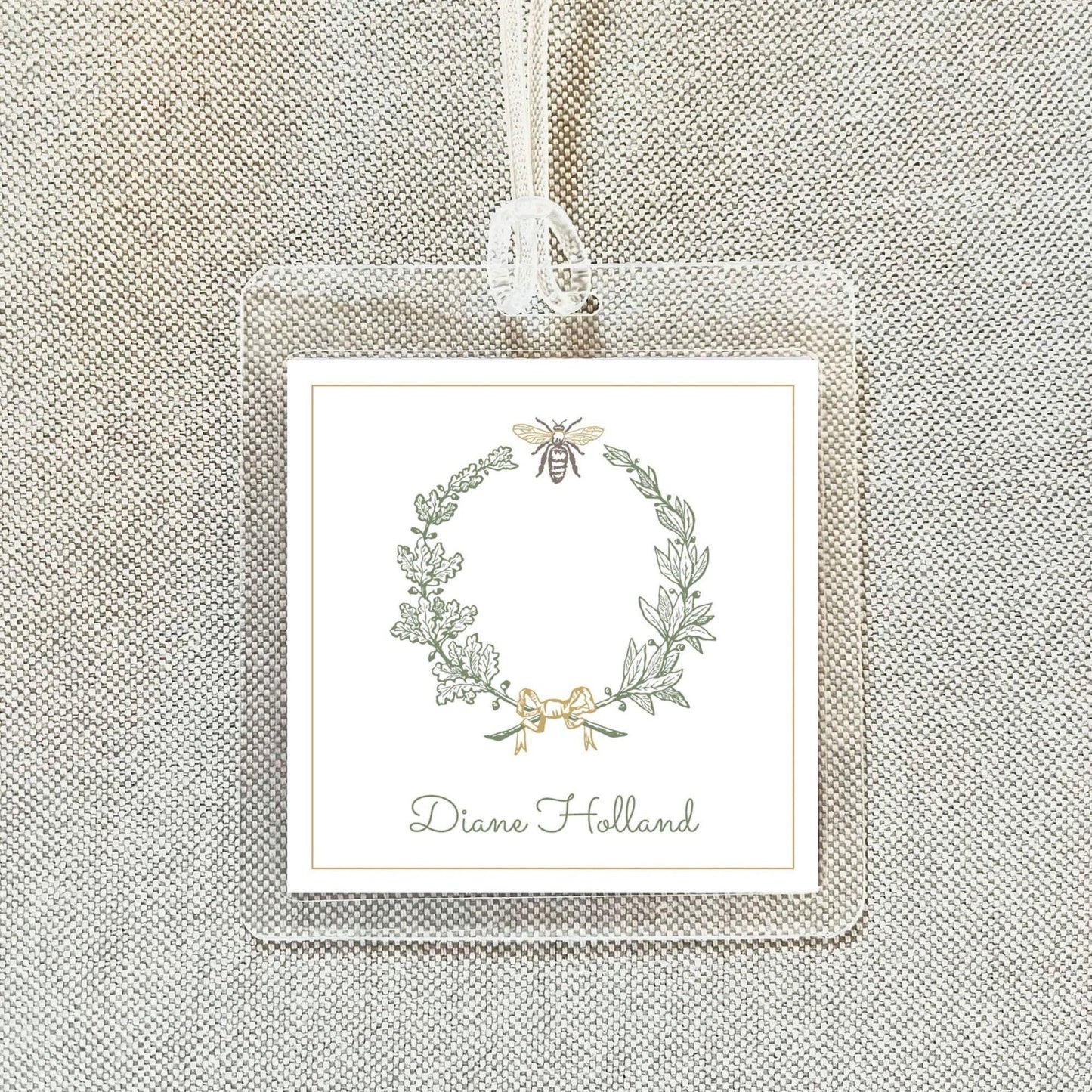 A square white label with a wreath design and a bee on it, hanging on a beige fabric background.