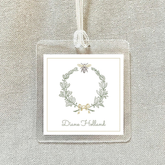 A square white label with a wreath design and a bee on it, hanging on a beige fabric background.