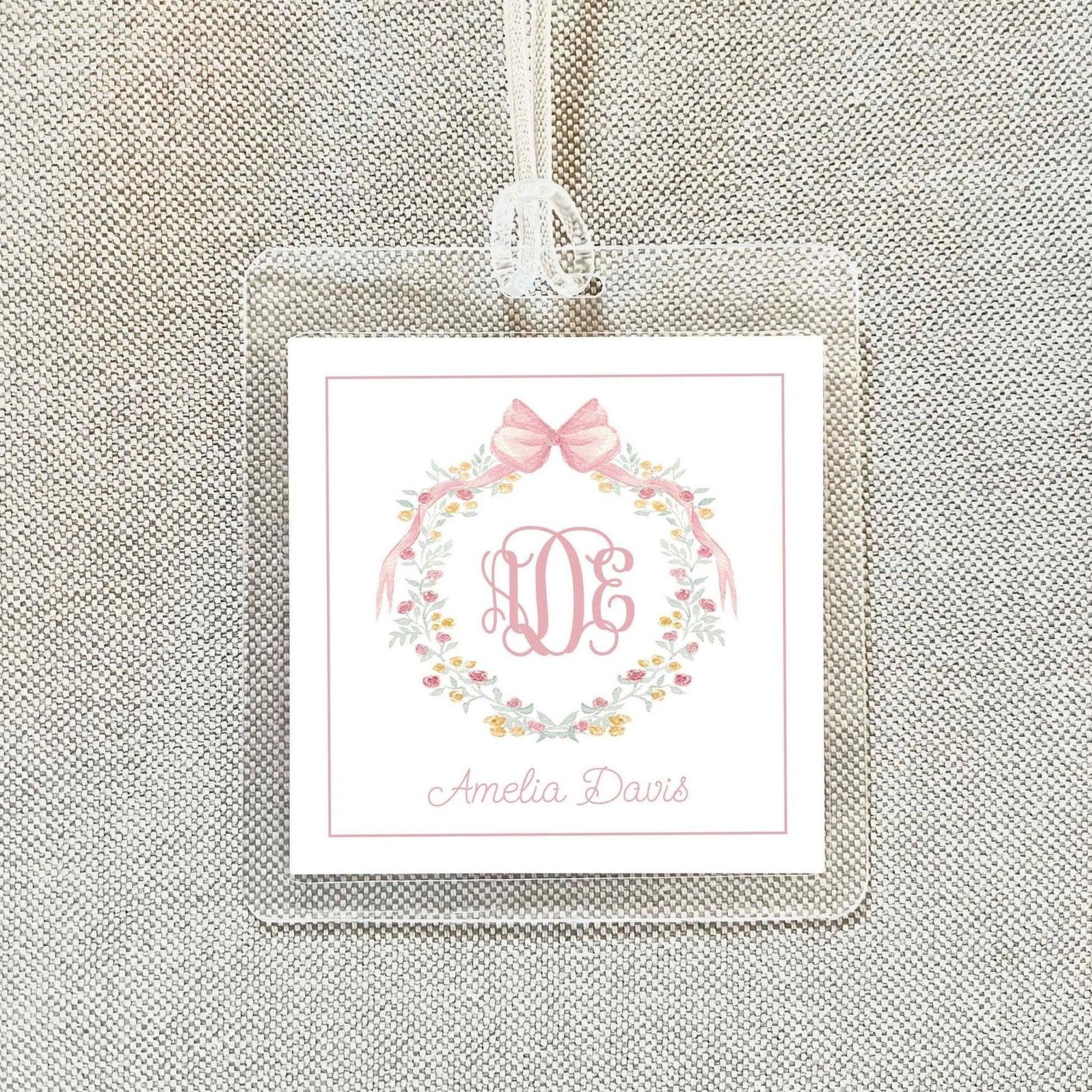 A clear plastic tag with a pink floral wreath and a monogrammed initials, hanging on a beige fabric background.