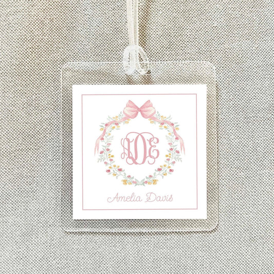 A clear plastic tag with a pink floral wreath and a monogrammed initials, hanging on a beige fabric background.