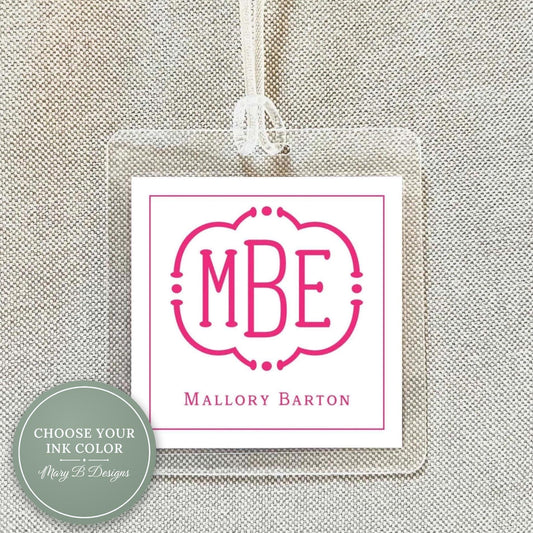 a clear plastic tag with a pink monogram logo and text, hanging on a beige fabric background.