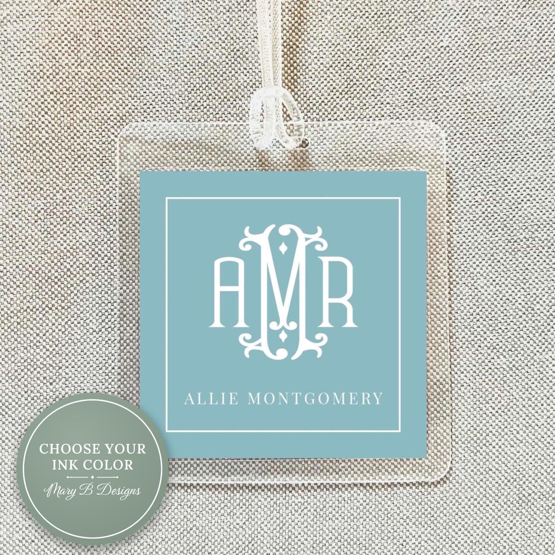 May include: Clear acrylic luggage tag with a light blue insert featuring a white monogram and the name "Allie Montgomery." The tag has a white string for attachment. The design is simple and elegant, suitable for travel.