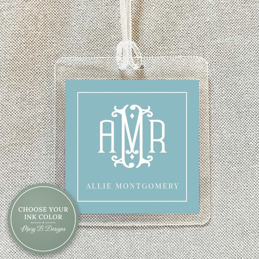 May include: Clear acrylic luggage tag with a light blue insert featuring a white monogram and the name "Allie Montgomery." The tag has a white string for attachment. The design is simple and elegant, suitable for travel.