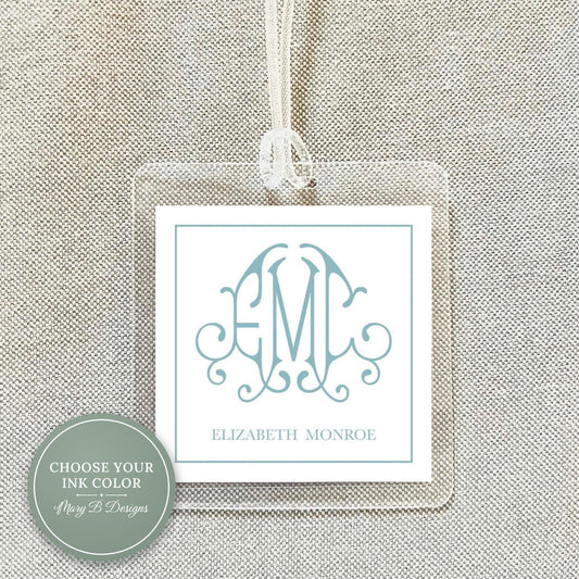 a clear plastic tag with a monogram design and text on it, hanging on a fabric surface.