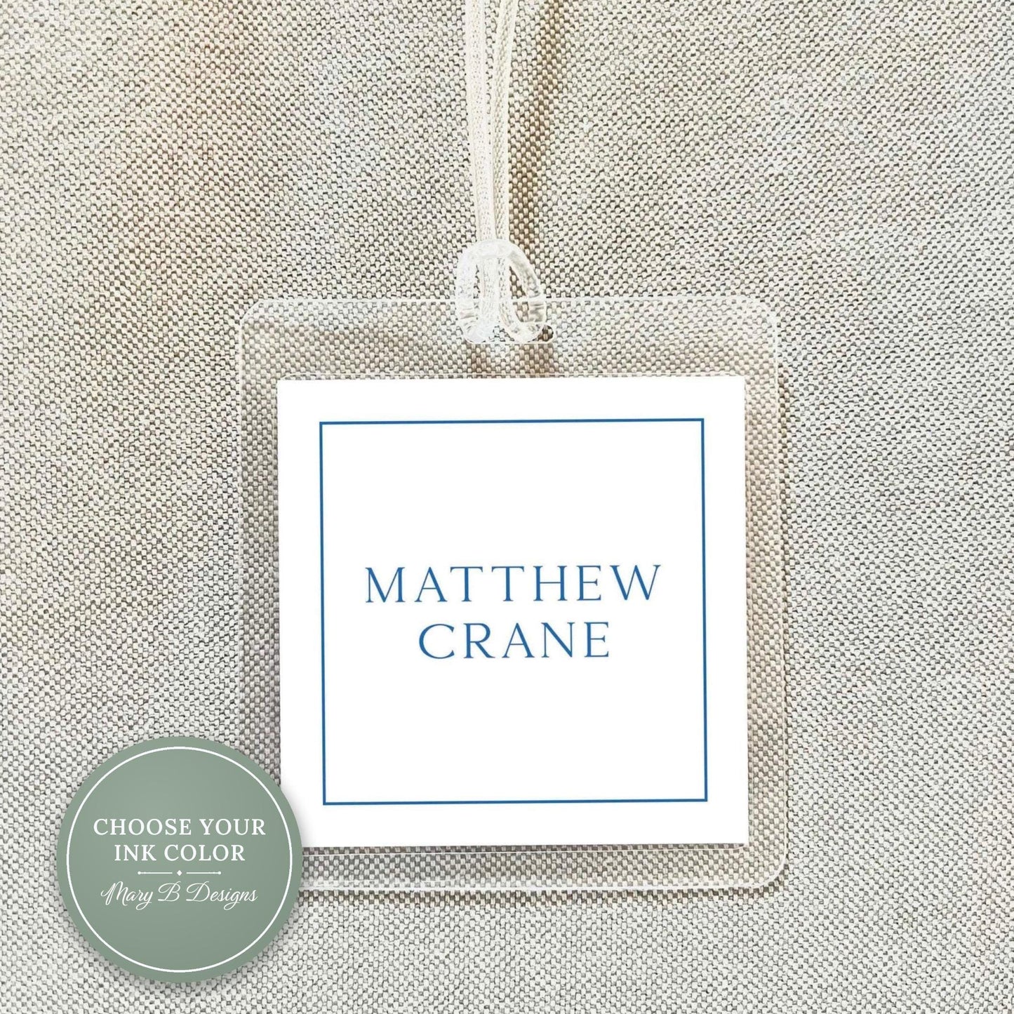 A clear plastic name tag with the name 'MATTHEW CRANE' printed on it, hanging on a fabric background.