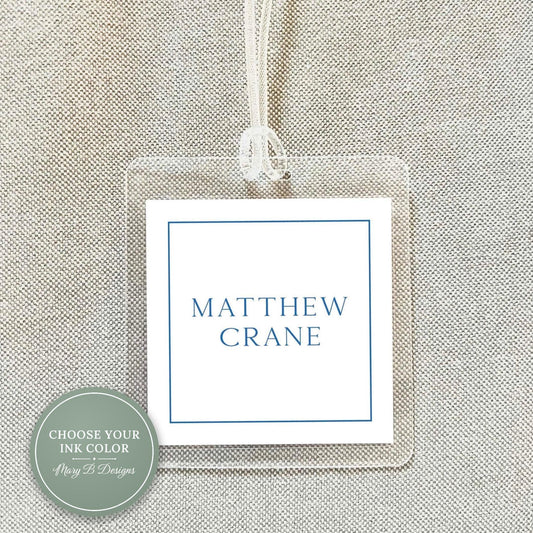 A clear plastic name tag with the name 'MATTHEW CRANE' printed on it, hanging on a fabric background.