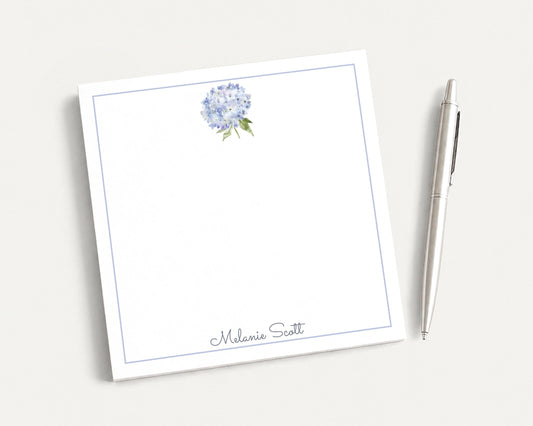 a white square notepad with a blue border, and a silver pen is placed next to it.