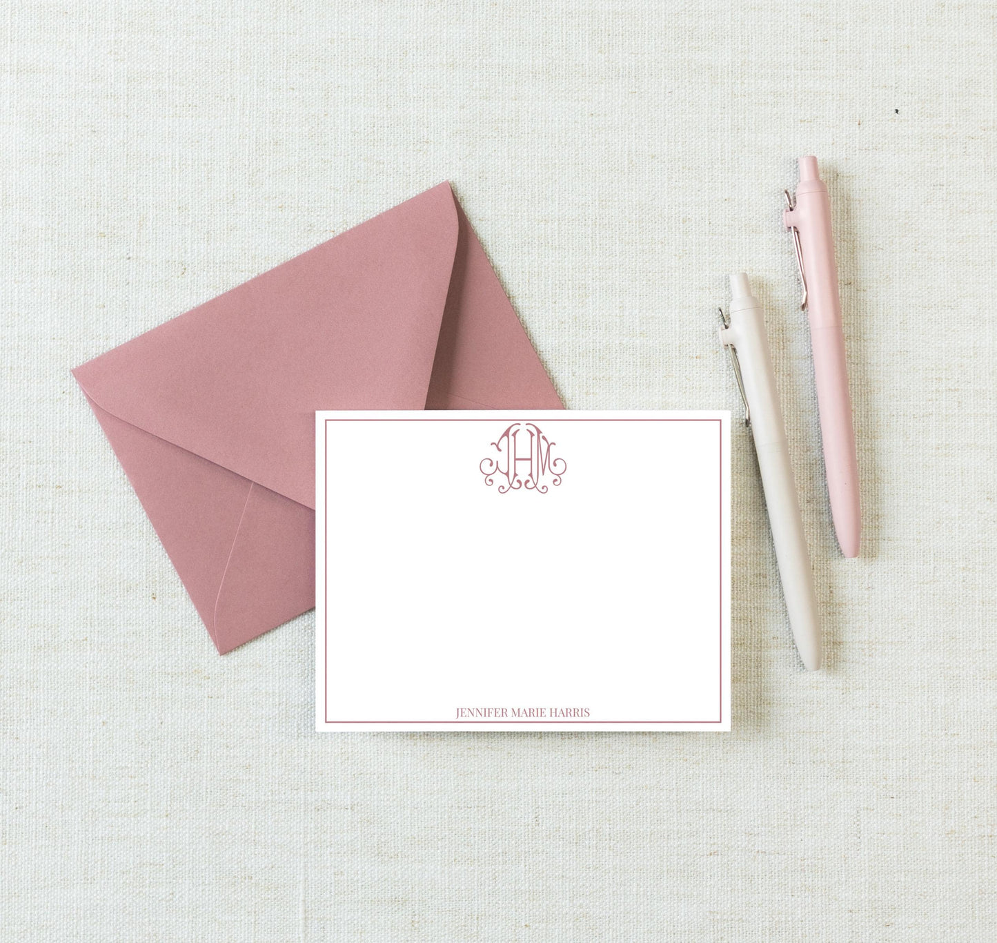 a pink envelope, a blank white card, and three pens arranged on a light-colored surface.