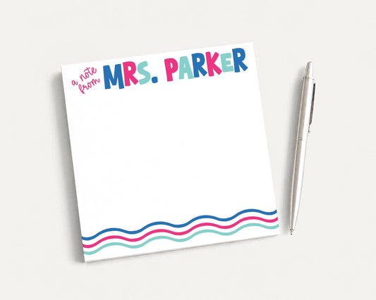 A colorful note pad with the name "Mrs. Parker" written on it, along with a pen, is placed on a white surface.