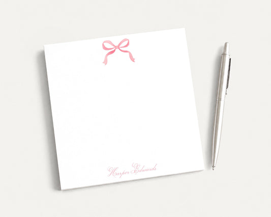 a white notepad with a pink bow and a pen on a plain white background.