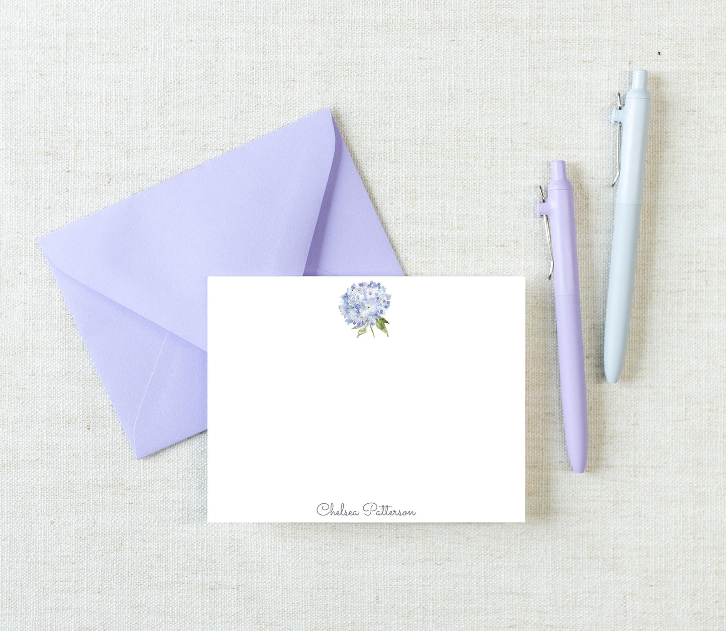 a purple envelope, a blank white card with a flower design, and two pens on a light-colored surface.