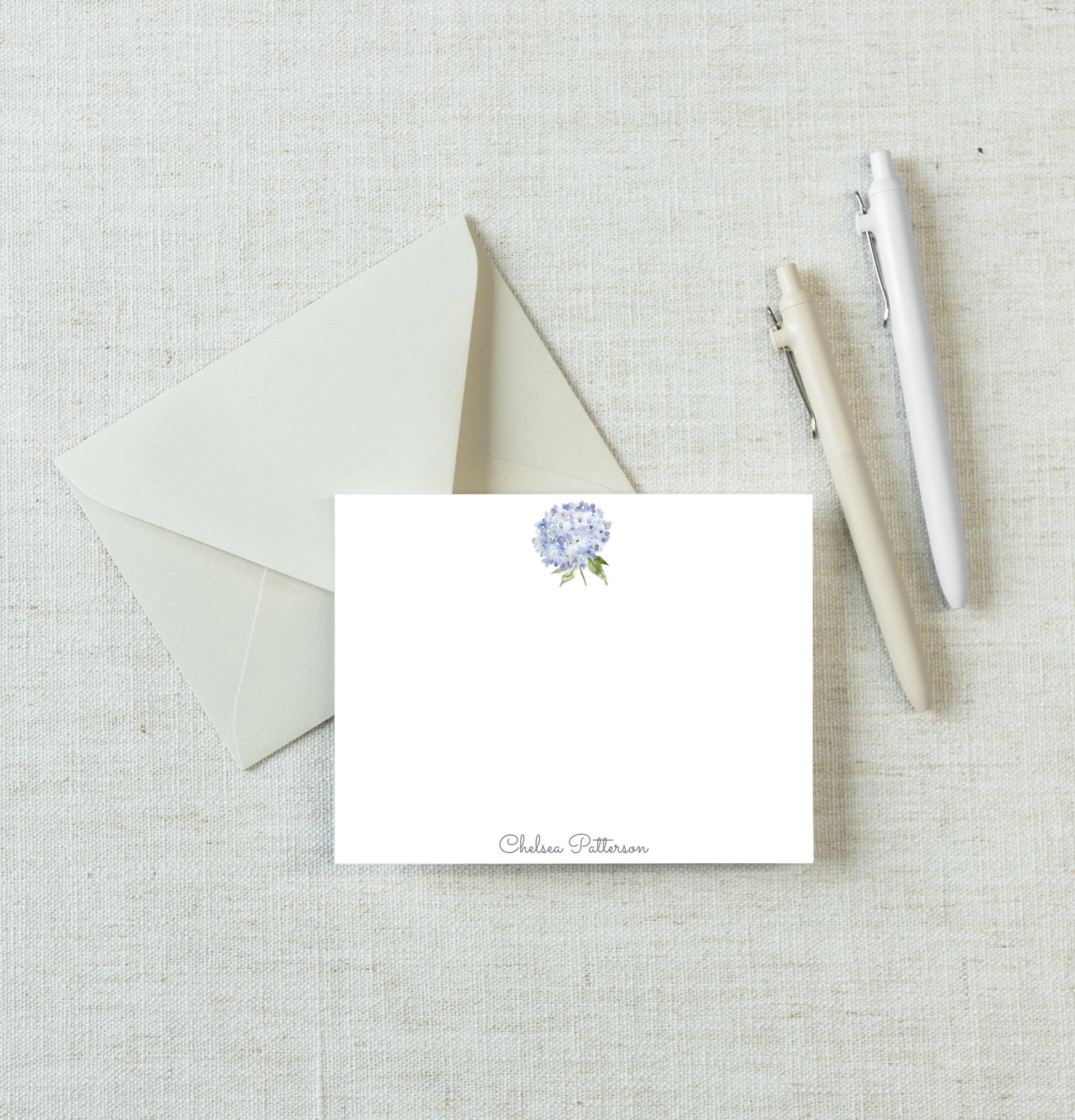 a white envelope with a blue flower design, a blank white card, and two pens on a light-colored surface.
