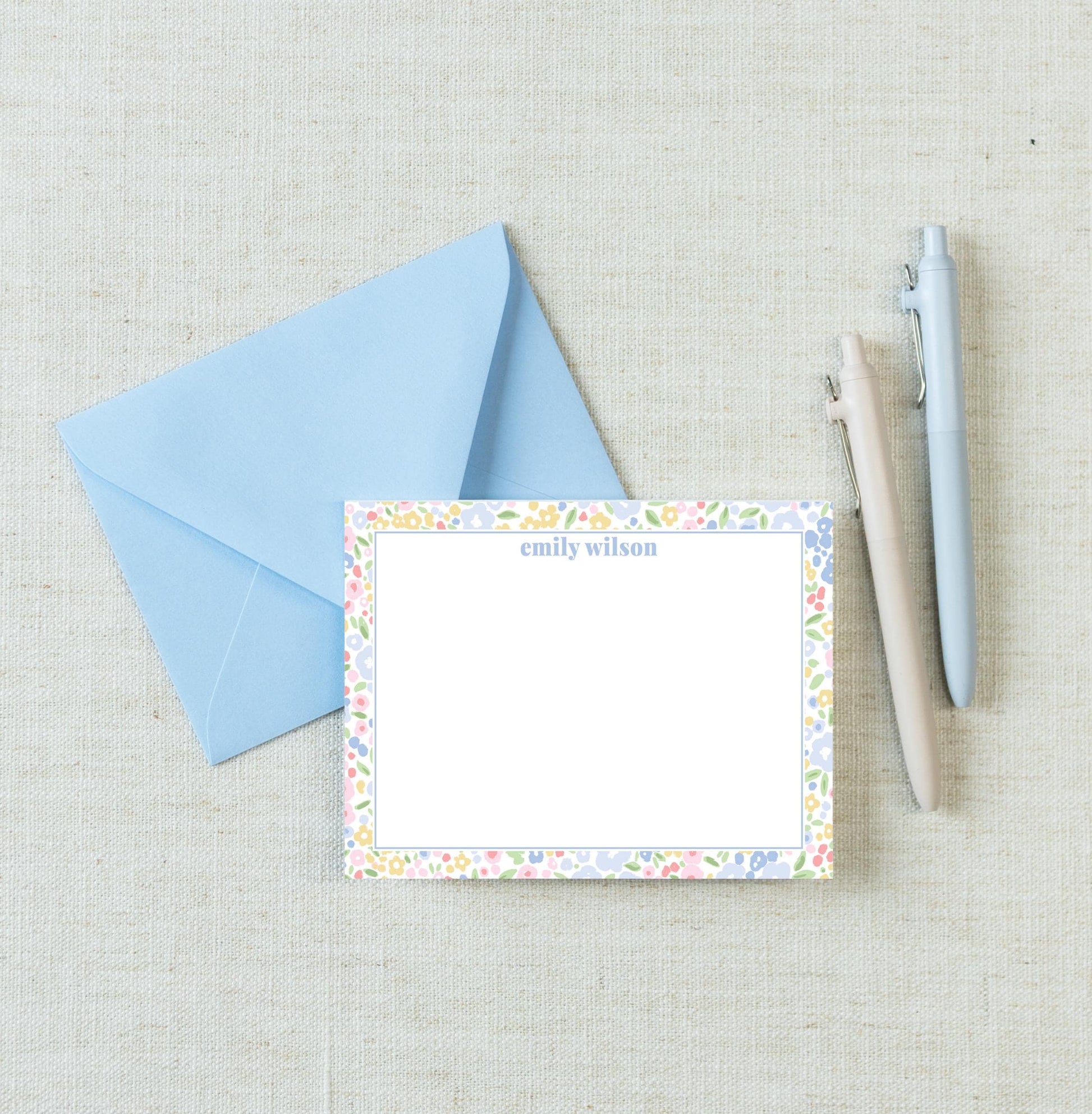 a blue envelope, a blank card with a floral border, and two pens on a beige background.
