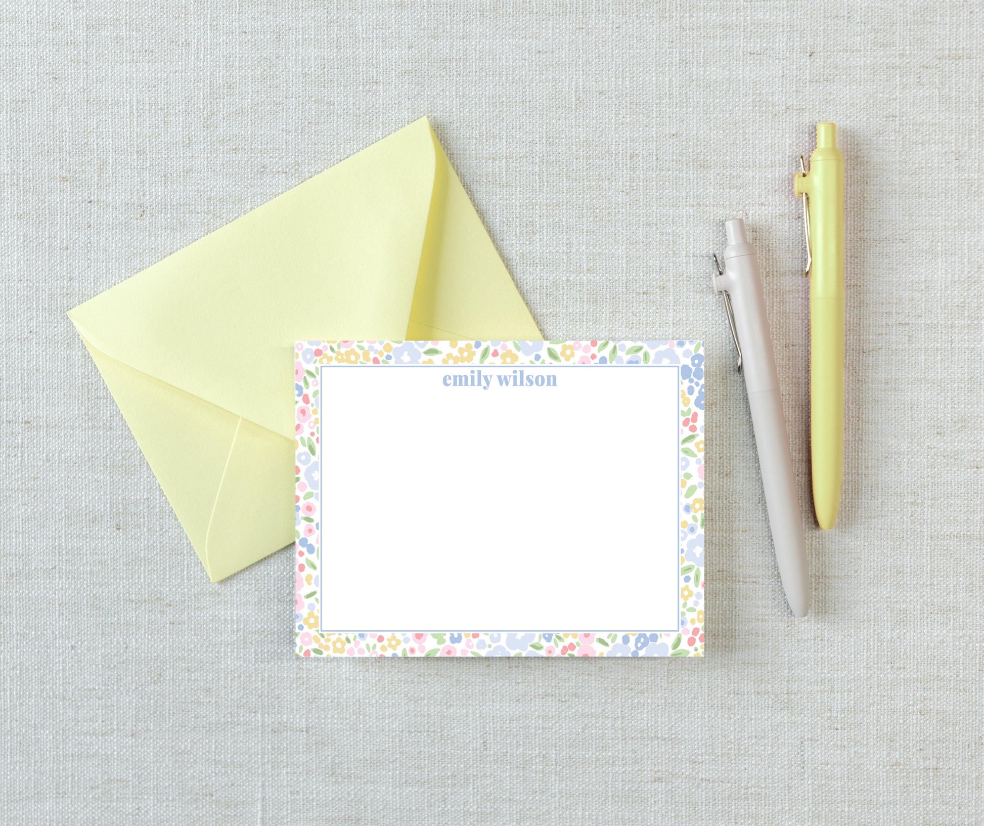 a yellow envelope, a blank card with a floral border, and two pens on a light-colored surface.