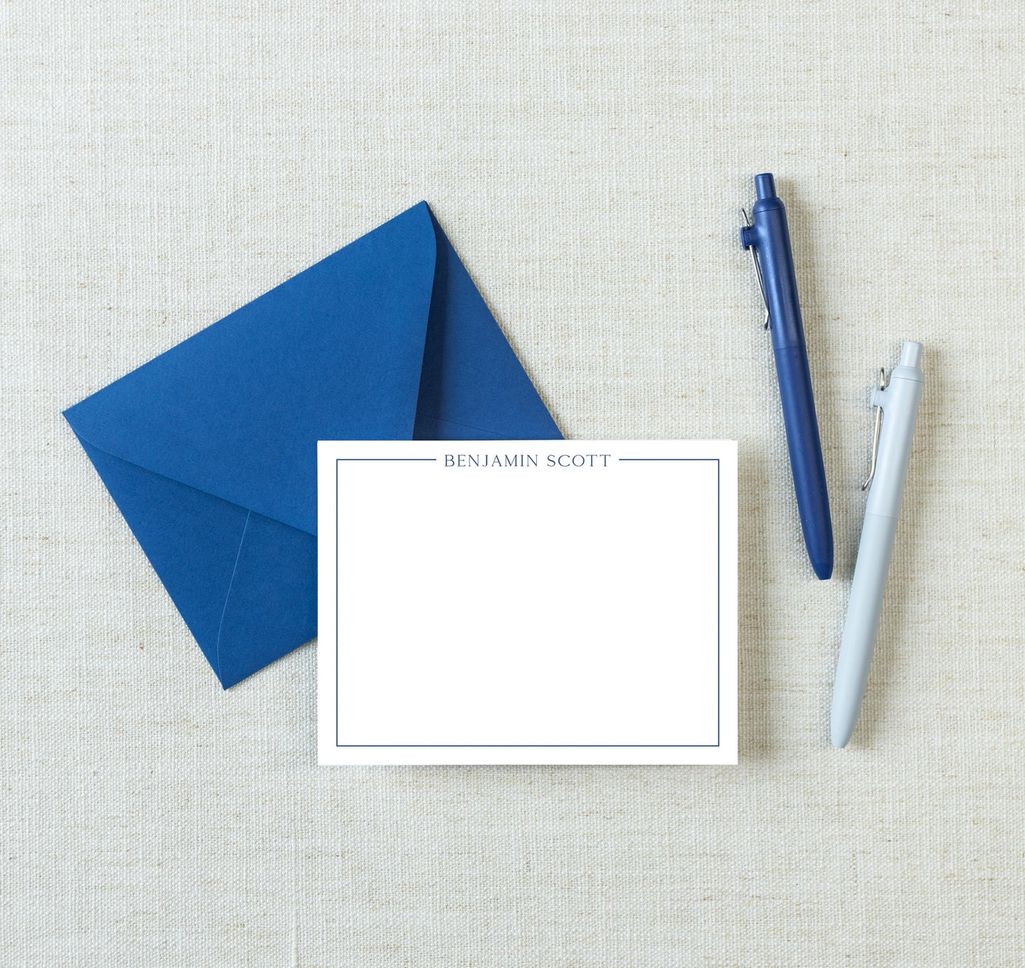 a blue envelope, a blank white card with the name "BENJAMIN SCOTT" written on it, and two pens, all placed on a beige background.