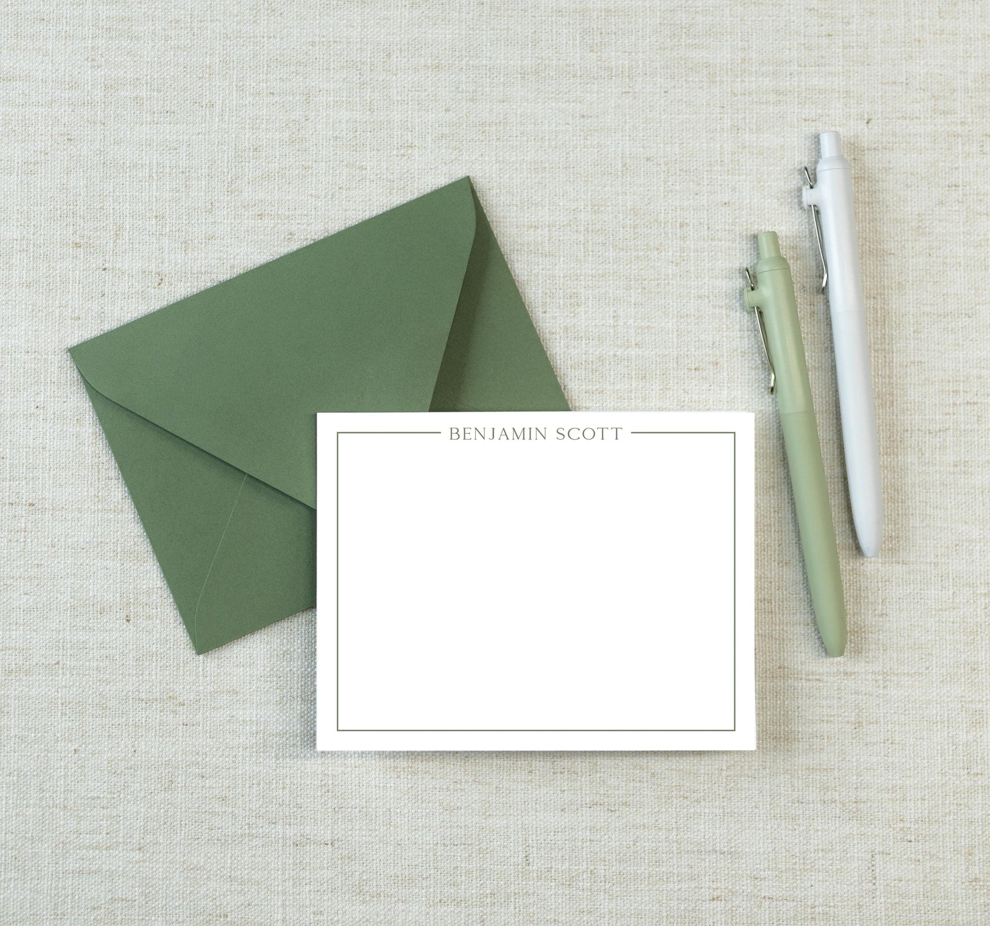 a green envelope, a blank white card with the name "Benjamin Scott" written on it, and two pens, all placed on a beige background.