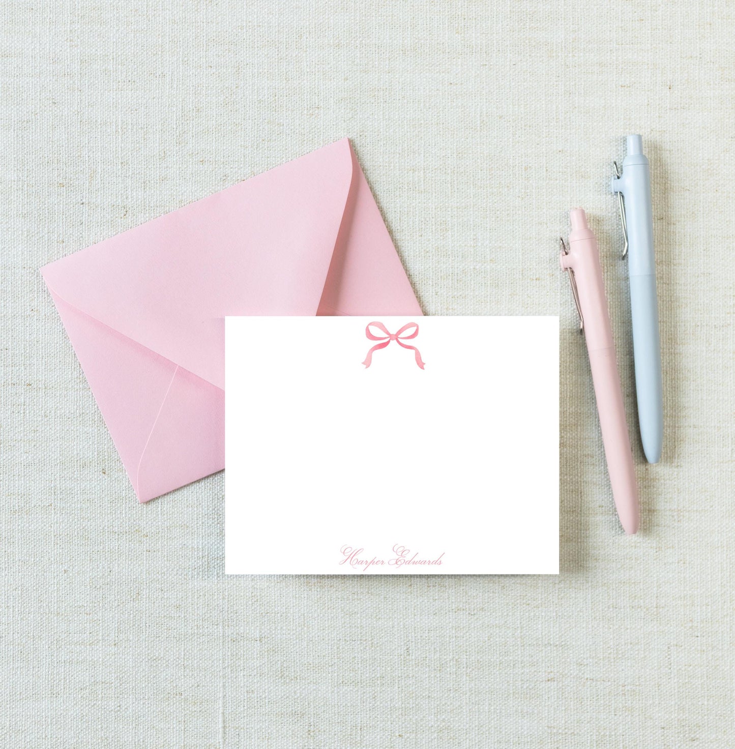 a pink envelope with a blank white card inside, along with two pens and a light-colored fabric background.