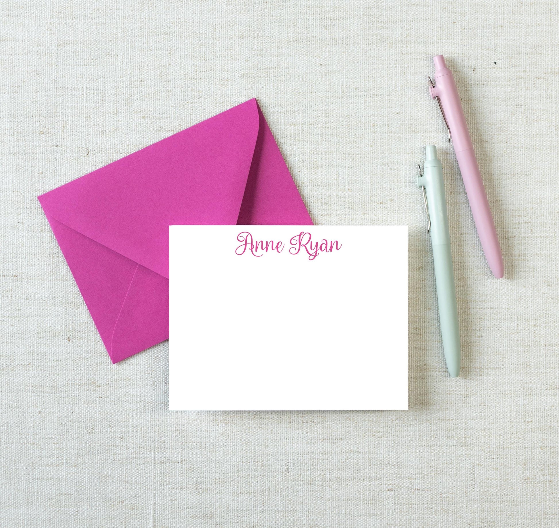 a pink envelope, a blank white note card with the name "Ana Ryan" written on it, and two pens, all placed on a light-colored fabric background.
