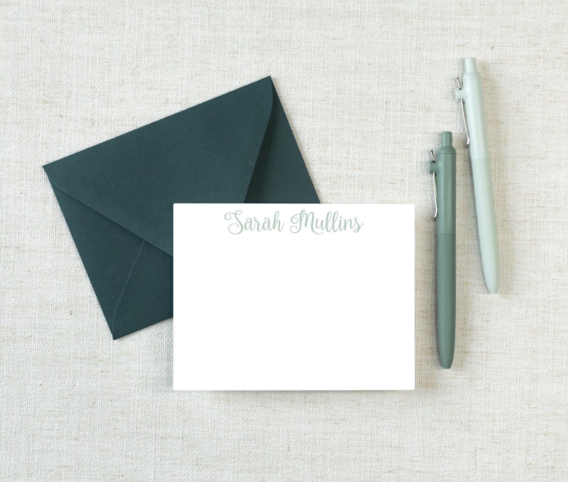 a green envelope with a blank white note card inside, along with two pens, all placed on a beige background.