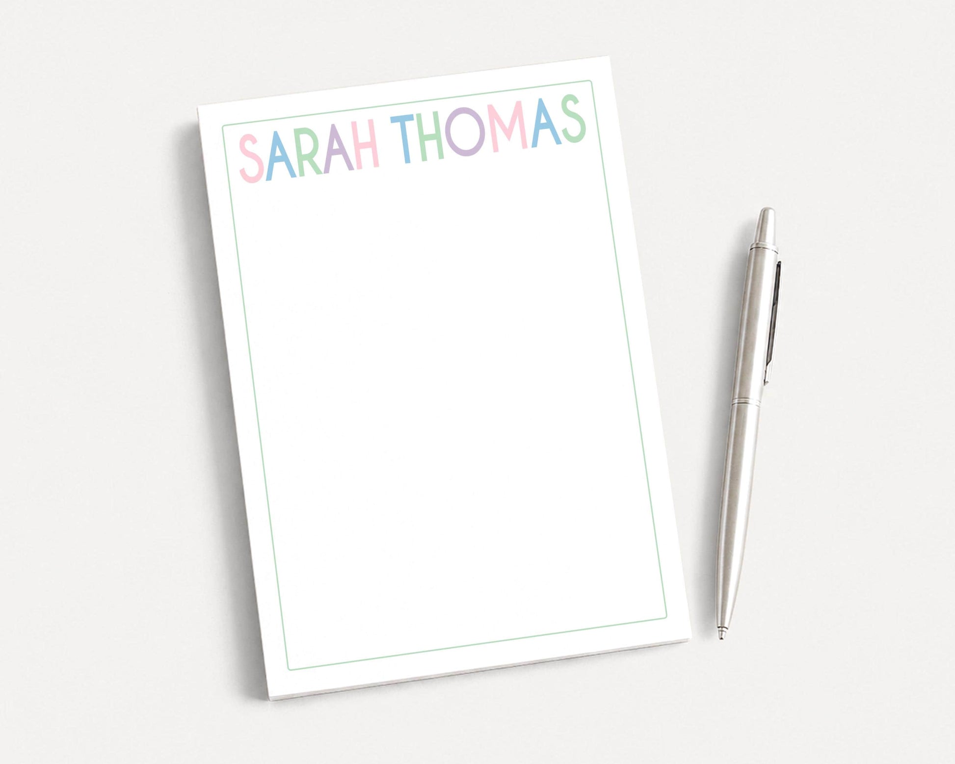 A blank white notepad with the name "SARAH THOMAS" written on it, along with a pen, is placed on a white surface.