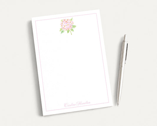 a blank white notepad with a floral design on the top left corner, accompanied by a silver pen on a plain white background.