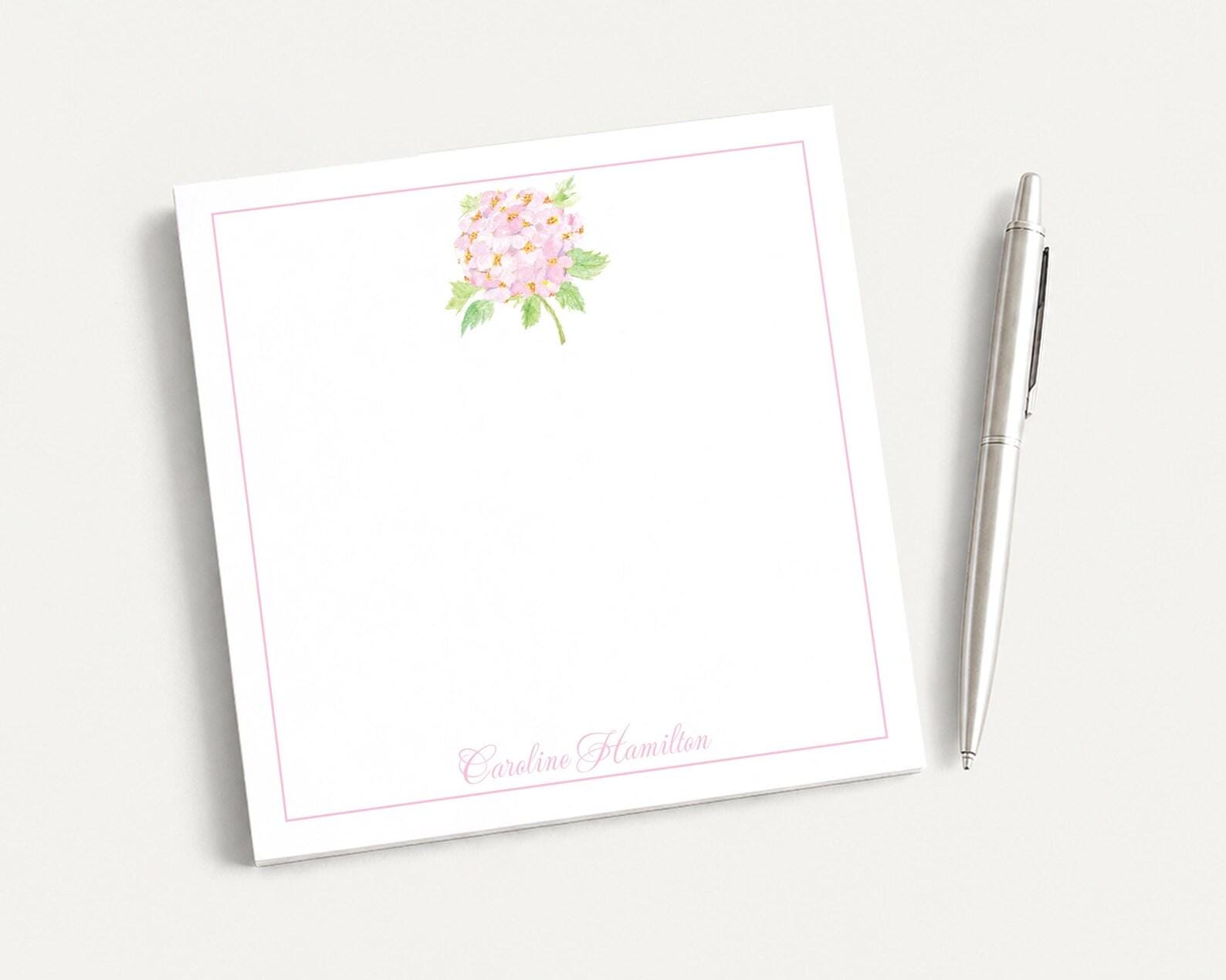 a white square notepad with a pink floral design and a pen placed next to it on a white surface.