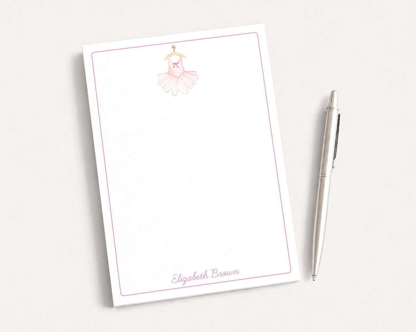 A blank white notepad with a pink border and a small illustration of a ballerina on the top left corner. Next to the notepad is a silver pen.
