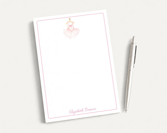 A blank white notepad with a pink border and a small illustration of a ballerina on the top left corner. Next to the notepad is a silver pen.
