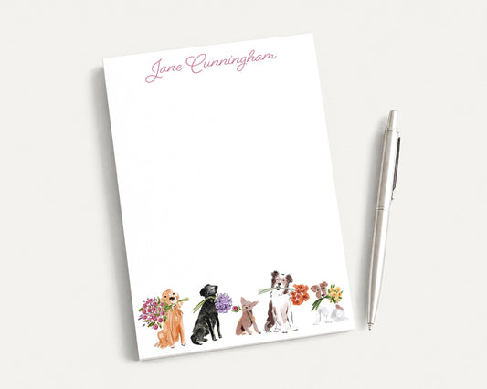 a white notepad with a watercolor illustration of five dogs of different breeds, each holding a flower. The notepad is accompanied by a pen on a white surface.