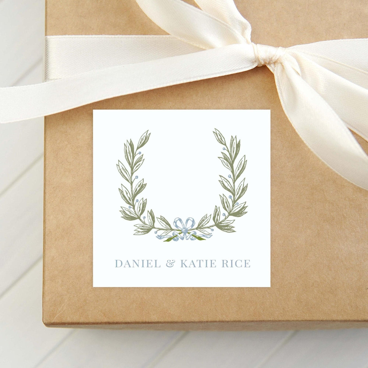 A brown gift box with a white ribbon tied around it, featuring a white label with a decorative wreath and the names "Daniel & Katie Rice" printed on it. __etsy:8651021647