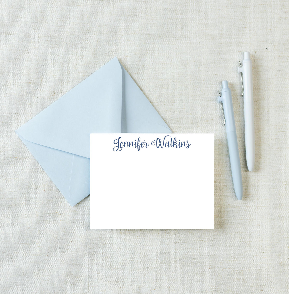 a light blue envelope with a blank white card inside, along with two pens on a light beige background. __etsy:8671806536