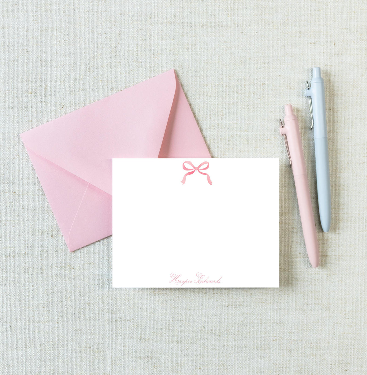 a pink envelope with a blank white card inside, along with two pens and a light-colored fabric background. __etsy:8719617619